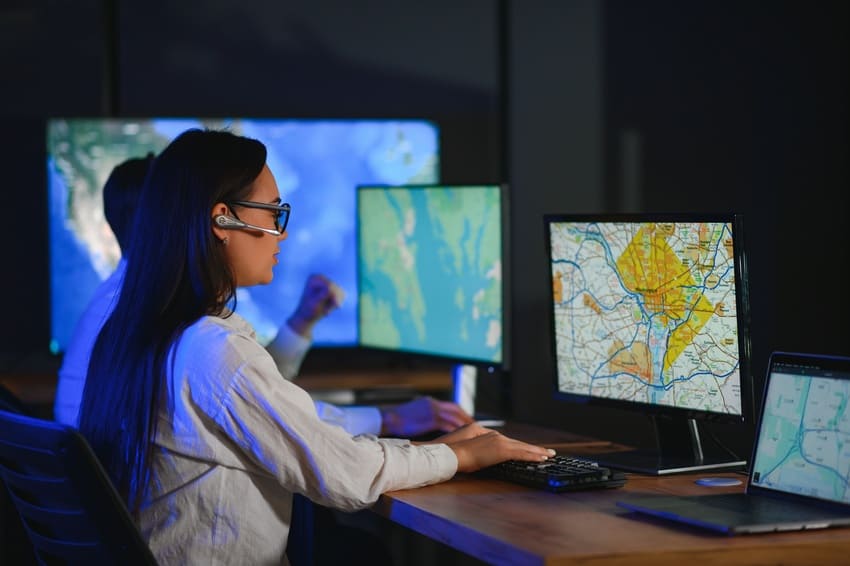 A person wearing a headset looks at digital maps on computer screens in a dark office environment.