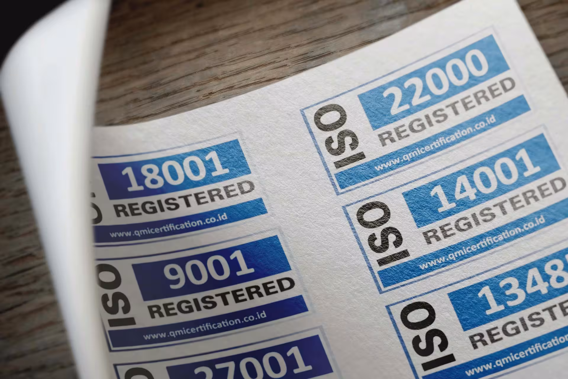 Close-up of several ISO certification labels, including ISO 9001, 14001, 18001, 22000, 27001, and 13485, displayed on a sheet of stickers on a wooden surface.